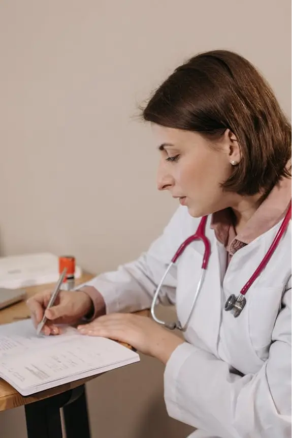 Female doctor in white coat with stethoscope writing notes on a clipboard at a desk.