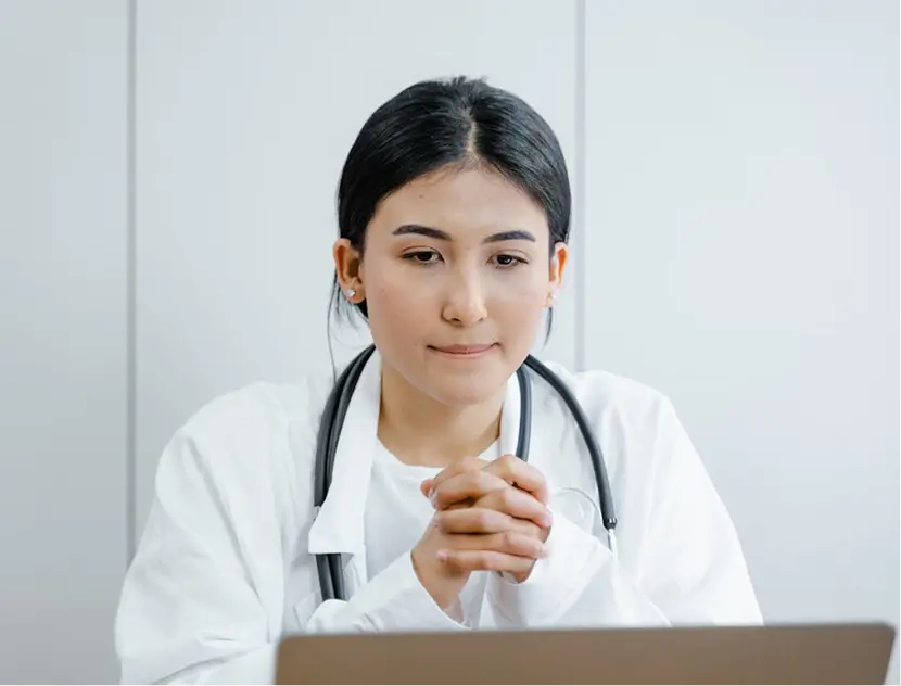 Female doctor with dark hair in a white coat and stethoscope looking thoughtfully at a laptop.