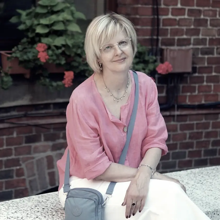Middle-aged woman with short blonde hair and glasses smiling, wearing a pink blouse and white skirt, sitting on a stone bench with a gray crossbody bag, against a brick wall background with green plants and red flowers.