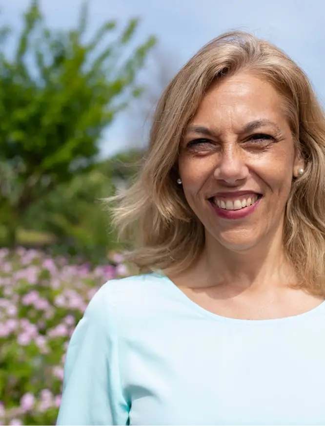 Smiling middle-aged woman with blonde hair wearing a light blue top standing outdoors with green trees and purple flowers in the background.