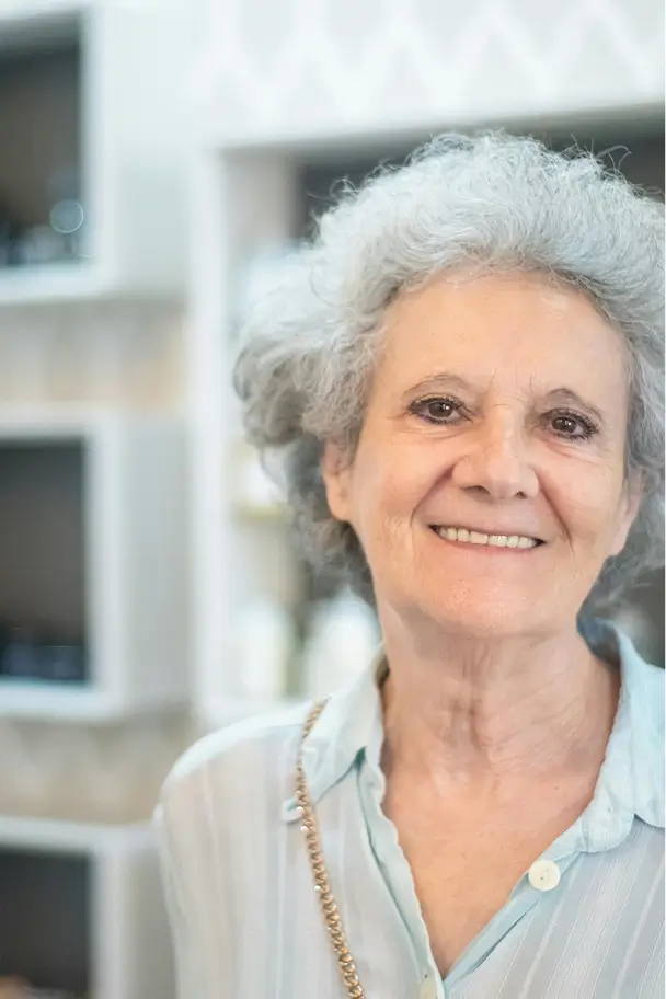 Smiling elderly woman with curly gray hair wearing a light blue shirt in an indoor setting.