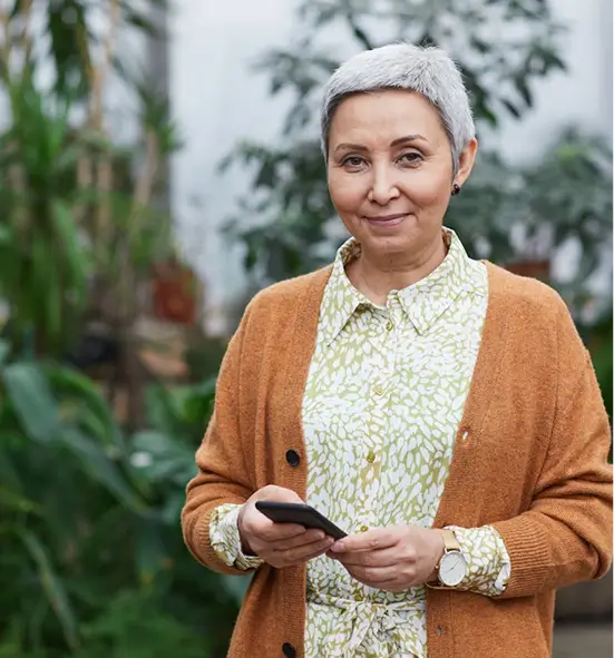 Smiling older woman with short gray hair wearing a patterned blouse and brown cardigan holding a smartphone in a green indoor garden.