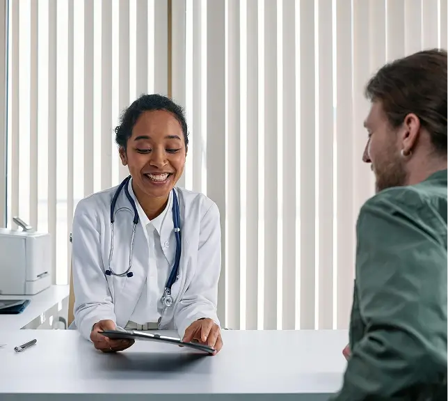 Smiling female doctor with a stethoscope showing a digital tablet to a male patient across a white desk in a well-lit room.