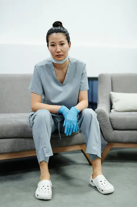 Tired healthcare worker in scrubs and blue gloves sitting on a couch with a mask under her chin.
