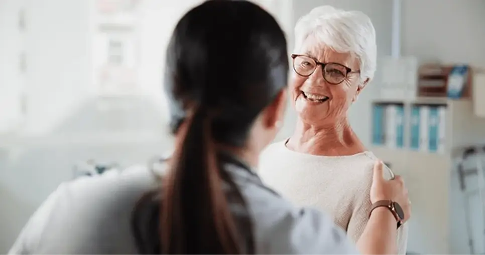 Smiling elderly woman with white hair and glasses holding hands with a person with dark hair in a healthcare setting.