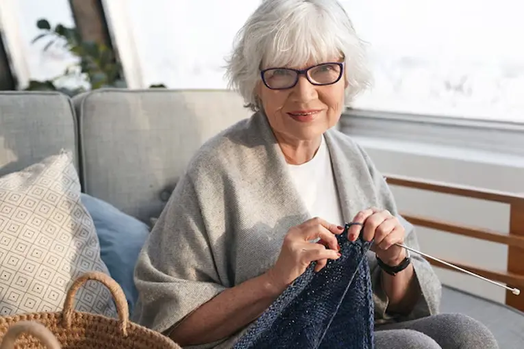 Elderly woman with white hair and glasses knitting a navy blue garment while sitting on a couch near a window.