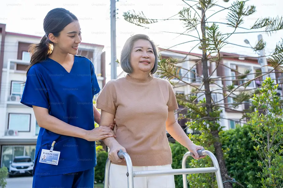 Young nurse in blue scrubs assisting elderly woman with a walker outside near residential buildings.