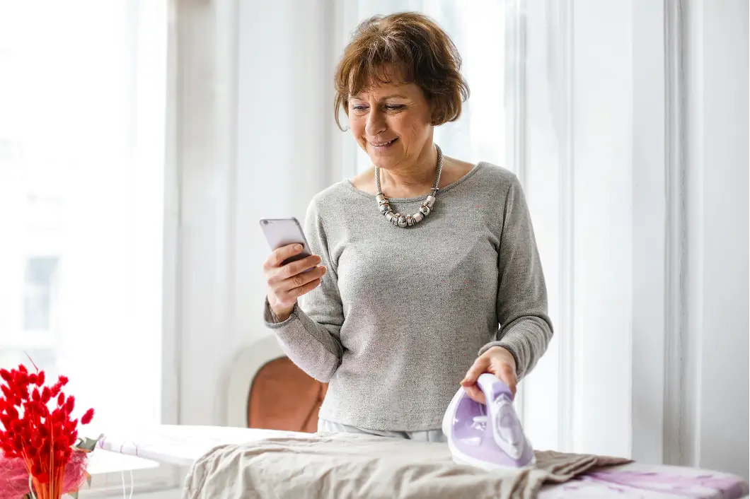 Middle-aged woman smiling while ironing clothes and looking at her smartphone in a bright room.