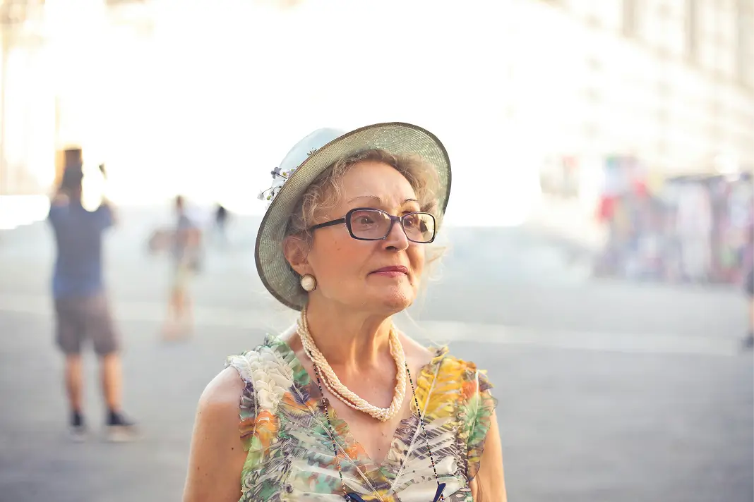 Elderly woman wearing glasses, a green hat, and a colorful floral dress standing outdoors in a bright urban area.