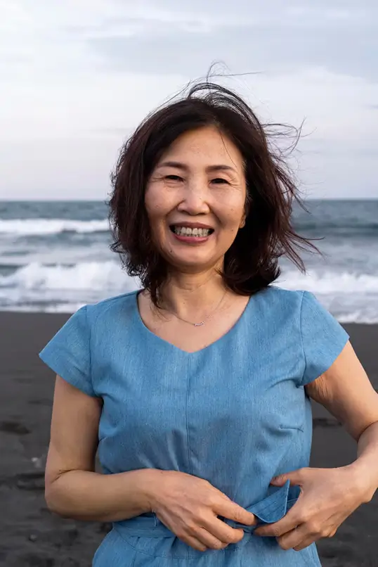 Smiling woman with shoulder-length dark hair wearing a blue dress standing on a beach with waves in the background.