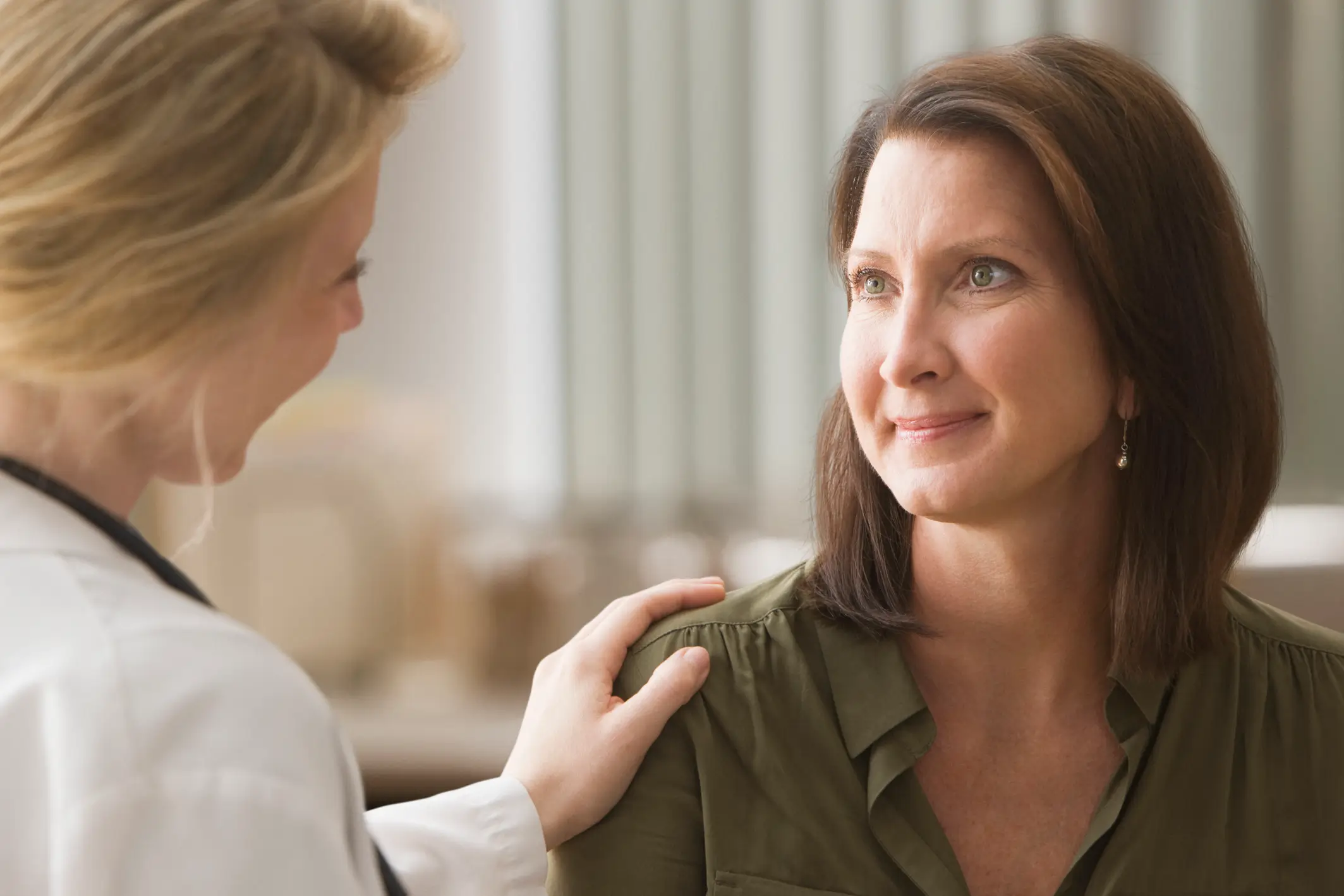 Doctor reassuring a middle-aged woman by gently placing a hand on her shoulder in a medical office.