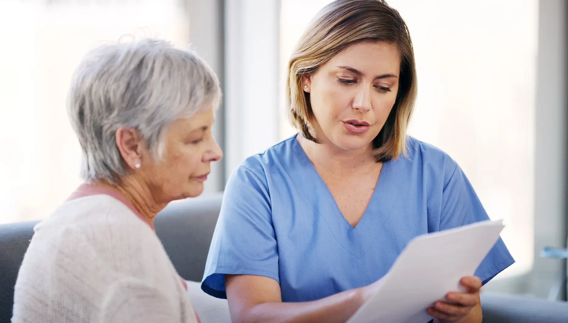 Healthcare professional in blue scrubs reviewing documents with an elderly woman in a light sweater.