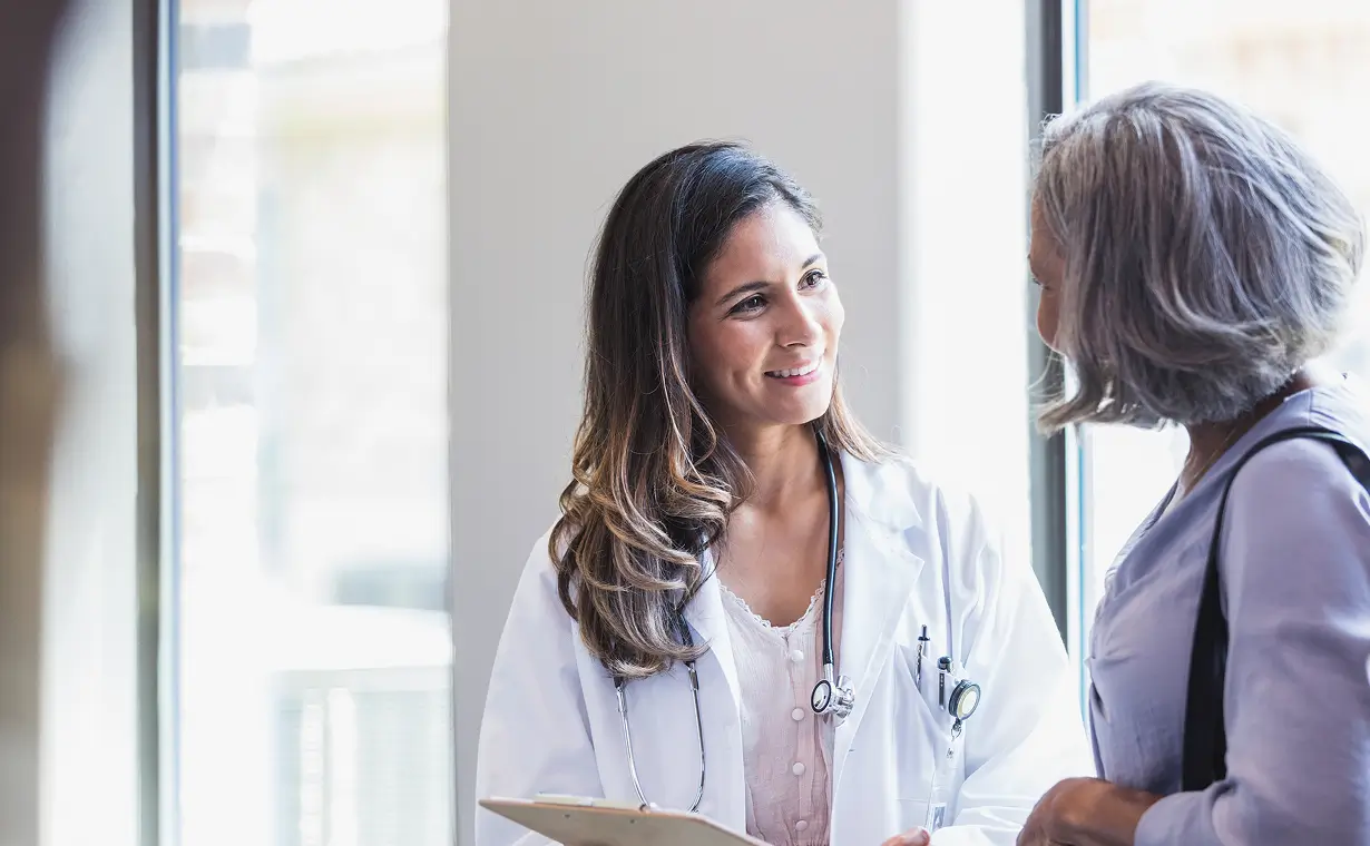Female doctor wearing a white coat and stethoscope smiling and talking with an older female patient by a window.