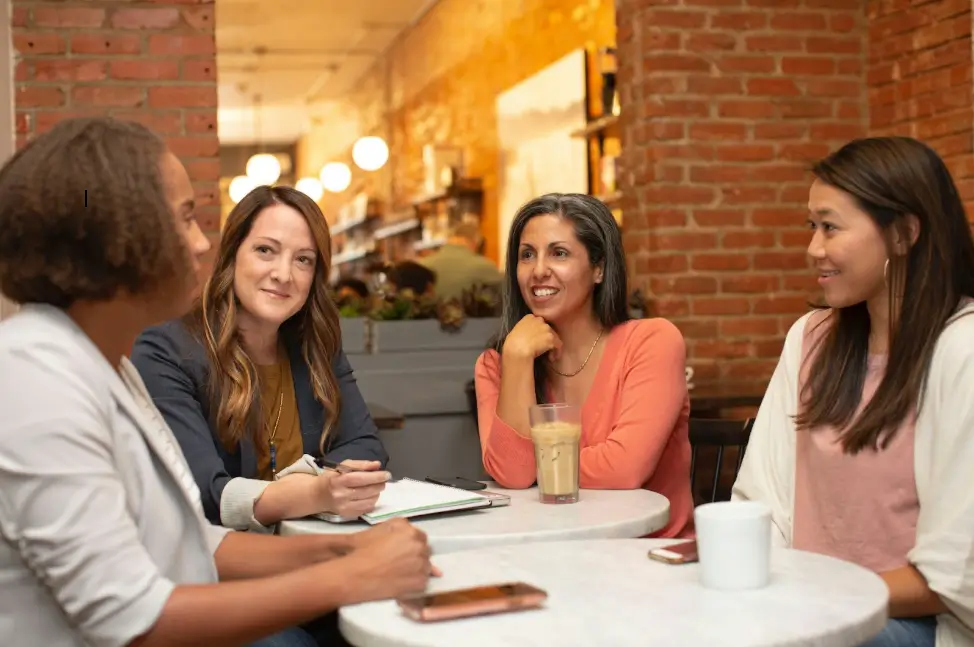 Four diverse women sitting around a table in a cozy café, engaged in conversation with notebooks, coffee, and a phone on the table.