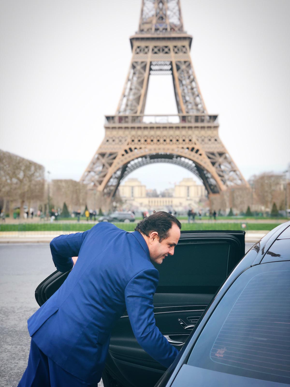 Homem de terno azul abrindo a porta traseira de um carro preto em frente à Torre Eiffel em Paris.