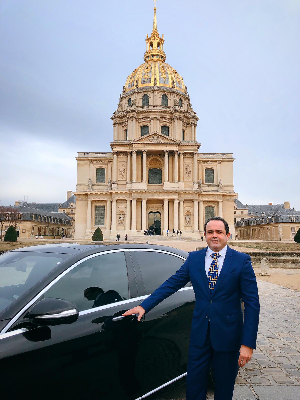 Homem de terno azul segurando a maçaneta de um carro preto em frente ao edifício com cúpula dourada Les Invalides em Paris.