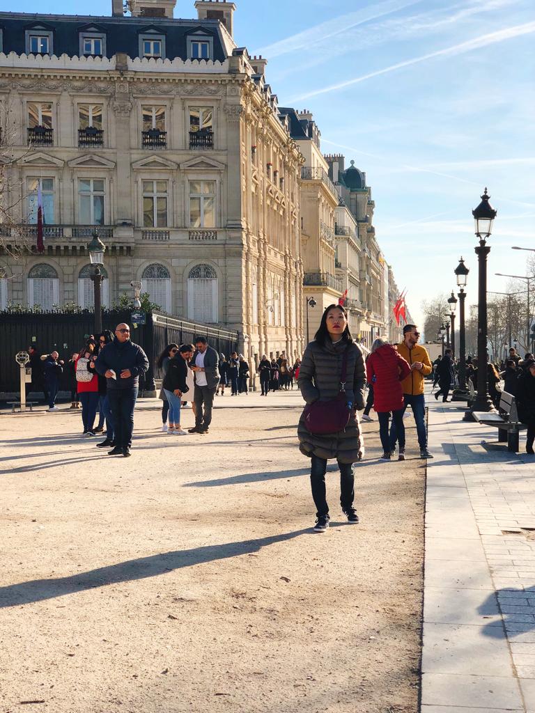 Pessoas andando em uma rua ensolarada com prédios históricos ao fundo, incluindo uma mulher usando casaco longo e bolsa roxa.