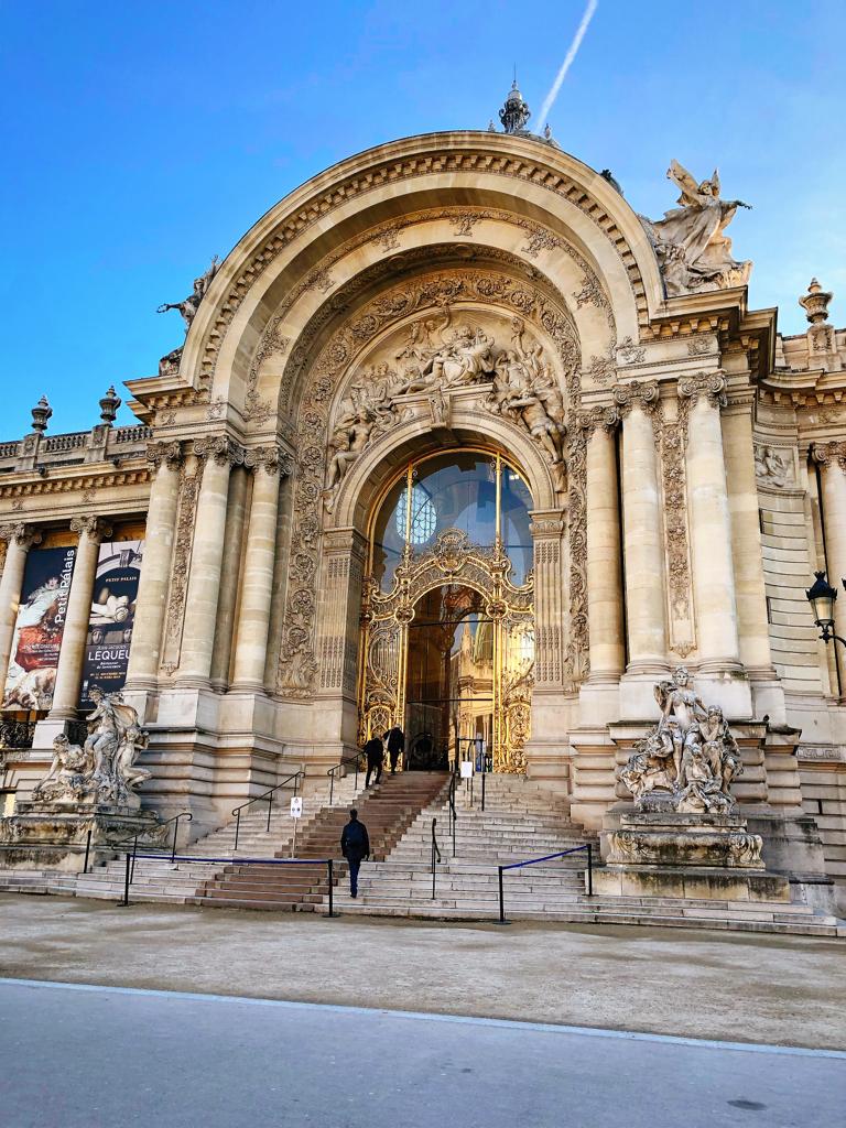 Entrada monumental do Petit Palais em Paris com grande arco ornamentado, escadaria e estátuas sob céu azul.