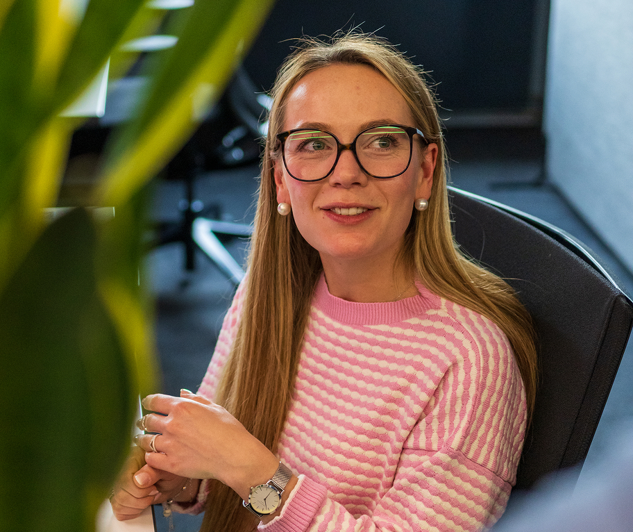 Frau mit langen blonden Haaren, Brille und rosa-weiß gestreiftem Pullover sitzt in Bürostuhl.