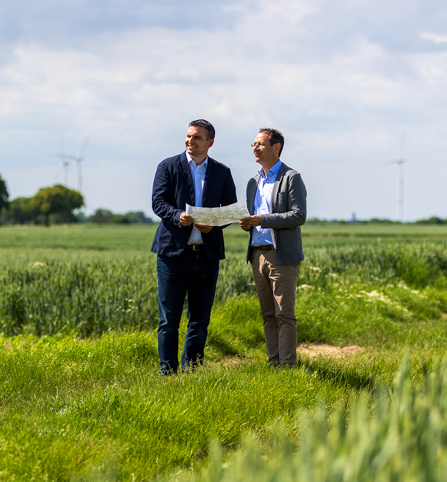 Zwei Männer in Anzügen stehen auf einem Feld und betrachten eine Karte bei windigen Windrädern im Hintergrund.