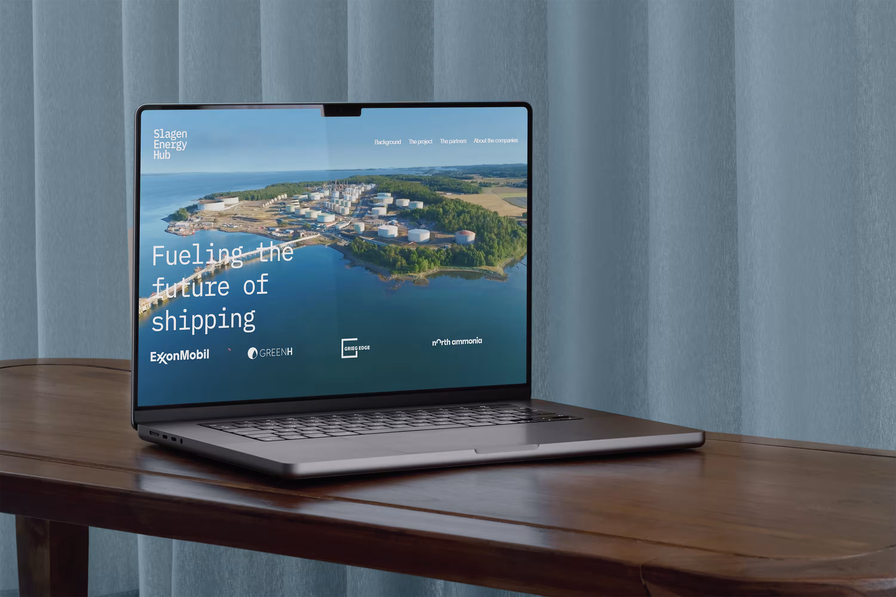 Laptop on wooden table displaying Slagen Energy Hub website with aerial view of industrial storage tanks near water.