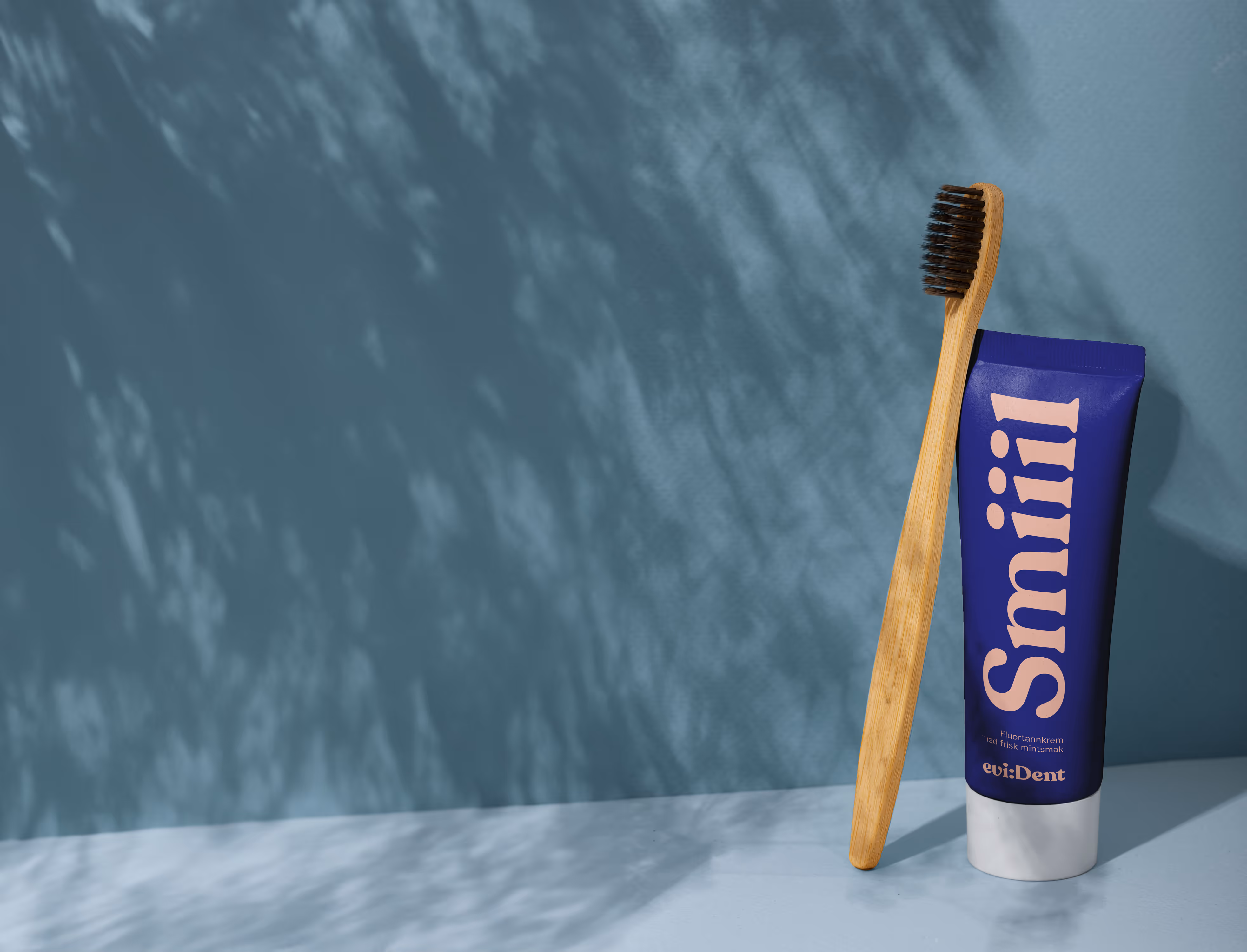 Wooden toothbrush leaning against a branded Evident's blue toothpaste tube on a blue surface with written "Smiiil" meaning "Smiiiile" in norwegian.
