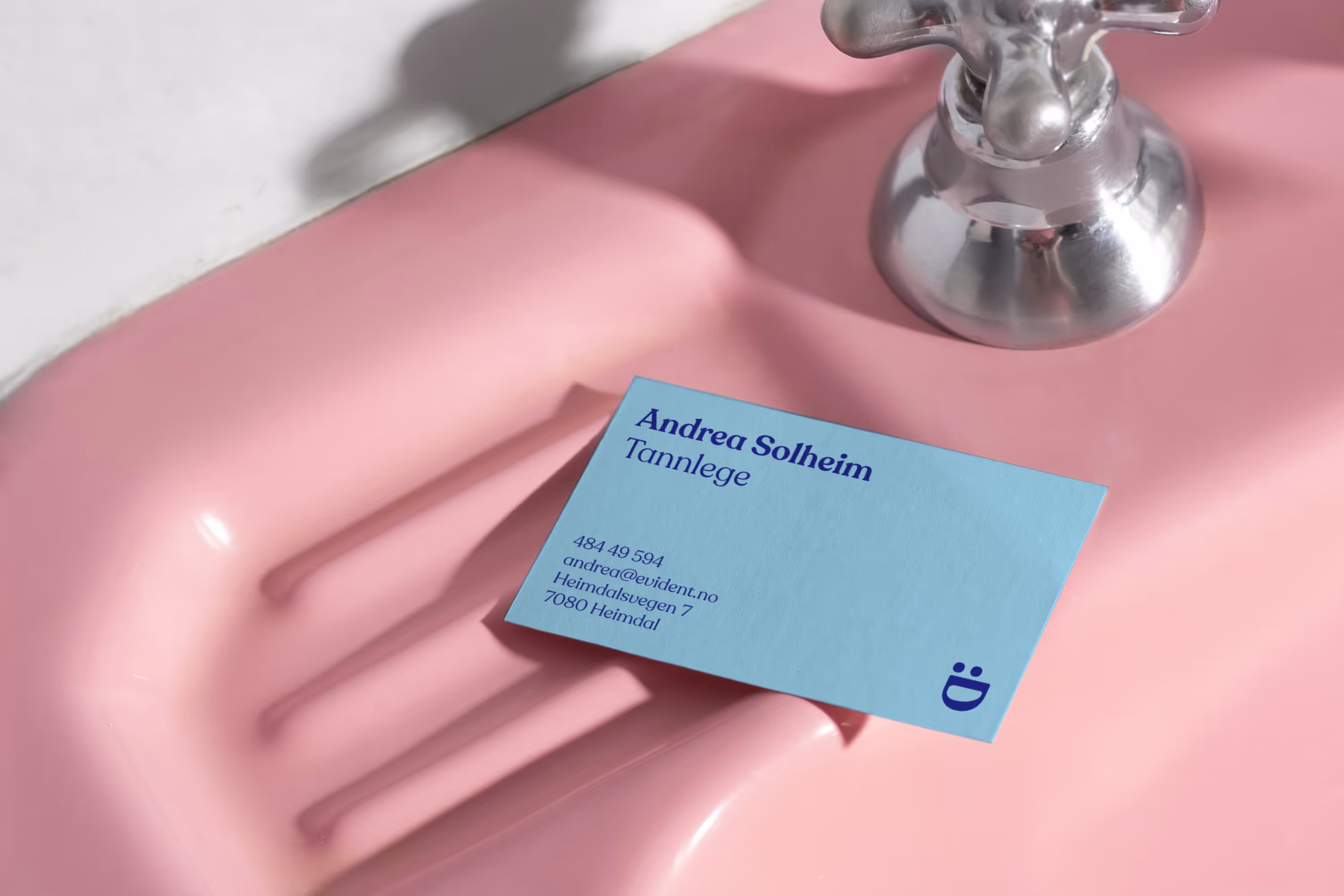 Blue business card for Andrea Solheim, a dentist from Evident, placed on a pink soap dish under a silver faucet.