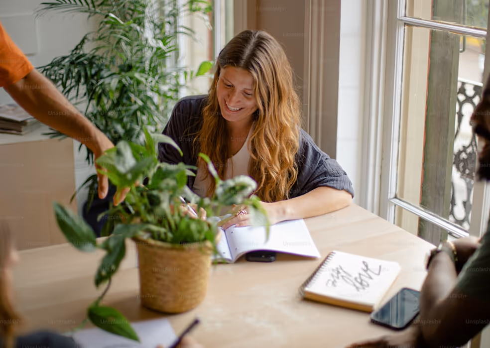 Smiling woman with long hair writing in a notebook at a wooden table surrounded by plants and other people.