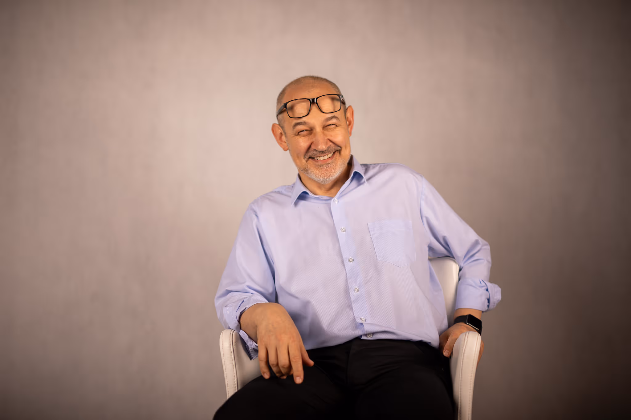 Usměvavý starší muž s brýlemi na hlavě sedí na bílé židli s šedým pozadím.