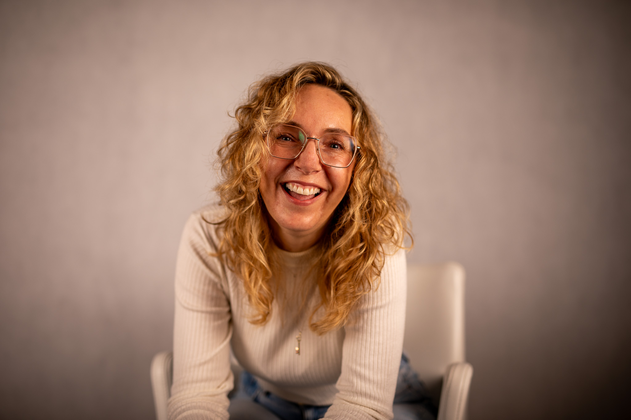 Usměvavá Kateřina Kurzová s kudrnatými blond vlasy a brýlemi sedí na židli před neutrálním pozadím.