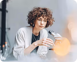 Woman with curly hair examining a sample through a microscope in a lab setting.
