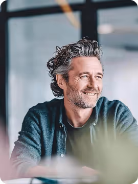 Smiling middle-aged man with curly gray hair and beard sitting indoors near a window.
