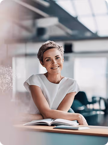 Smiling woman with short hair sitting at a table with an open book in a bright, modern office.