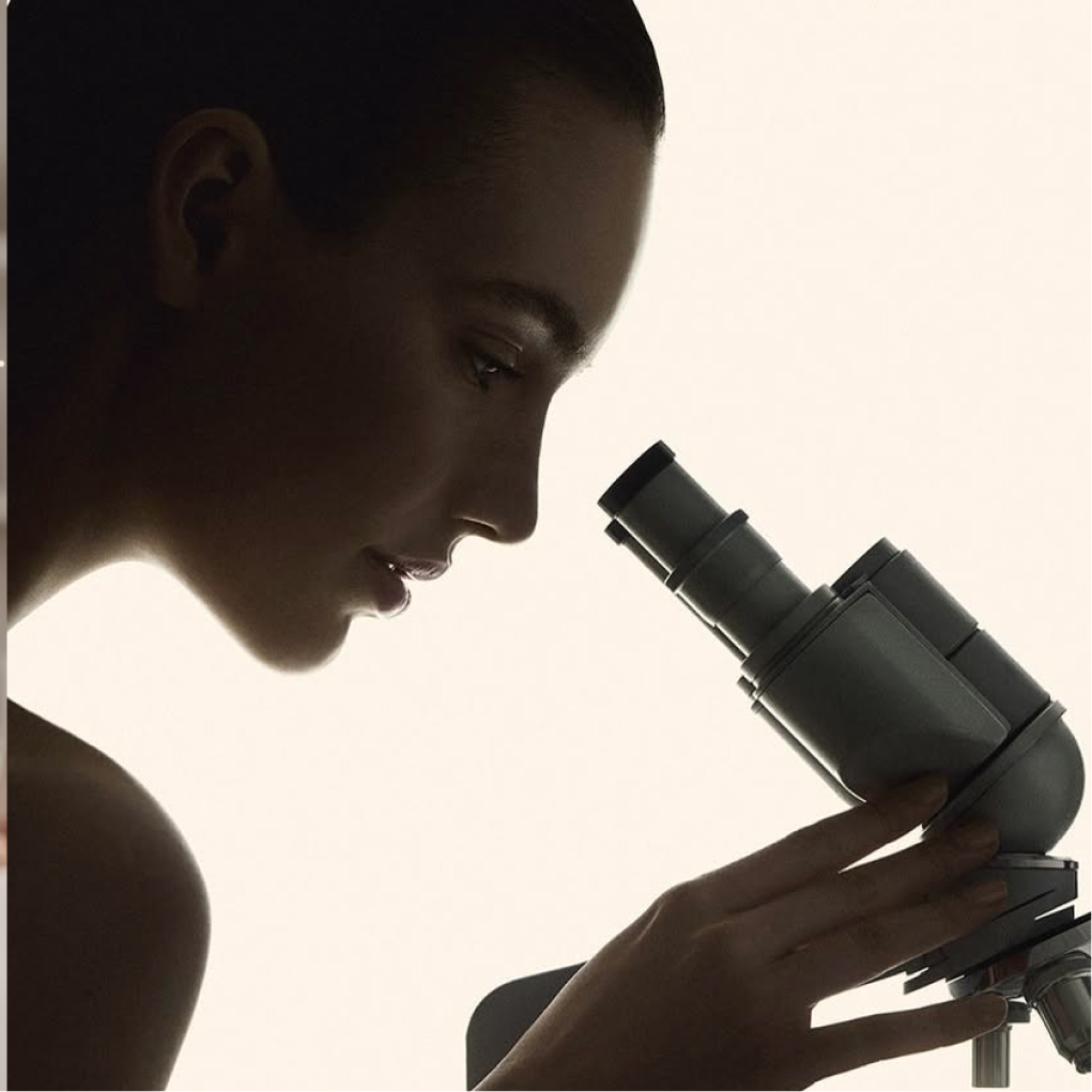 Side view of a woman looking through the eyepiece of a microscope against a light background.
