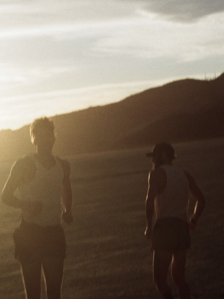 Two runners in tank tops and shorts jogging outdoors at sunset near hills.