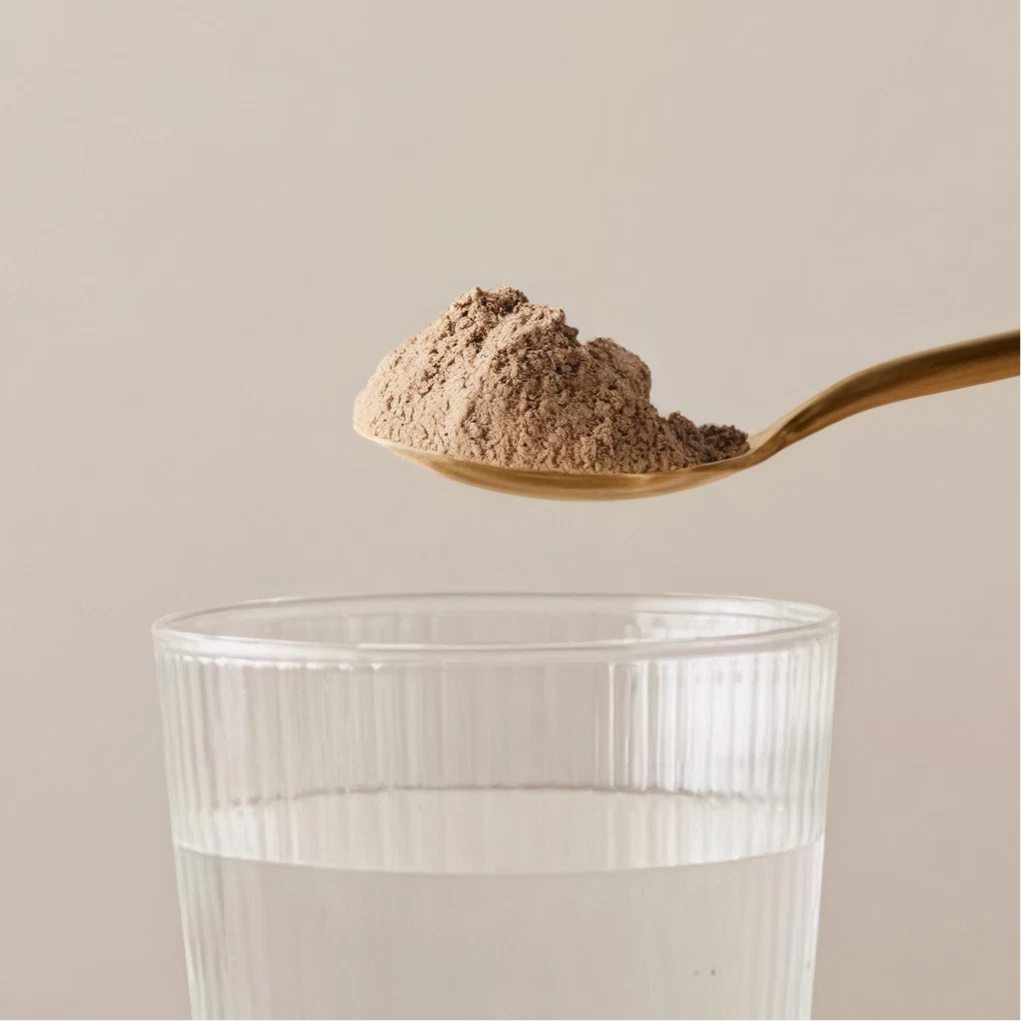 Spoon of light brown powder held above a clear glass of water against a plain background.
