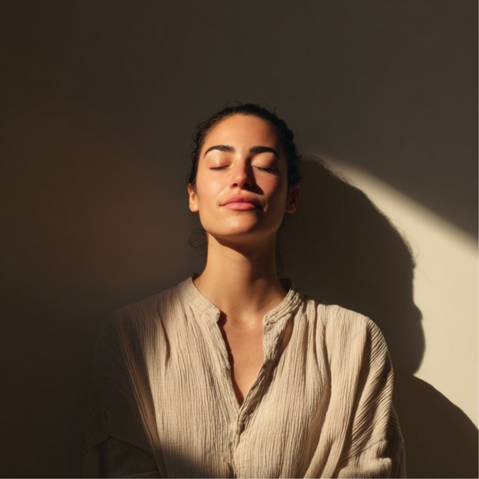 Woman with closed eyes standing in soft sunlight casting a shadow on a beige wall behind her.