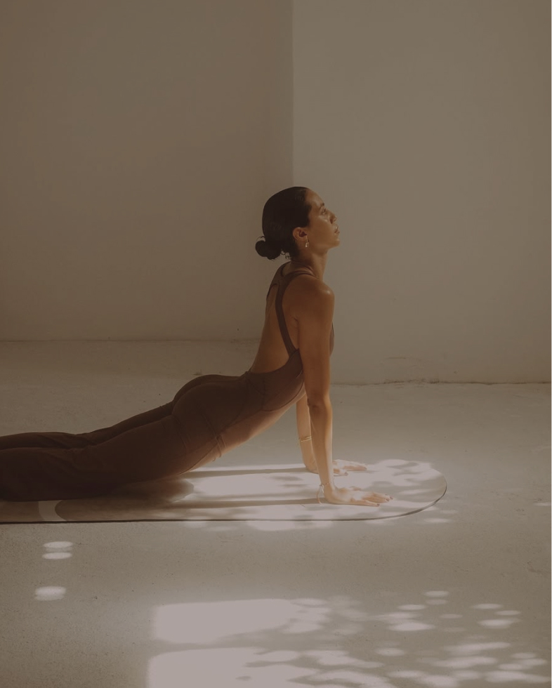 Woman in a dark outfit doing yoga cobra pose on a mat in soft sunlight inside a room.