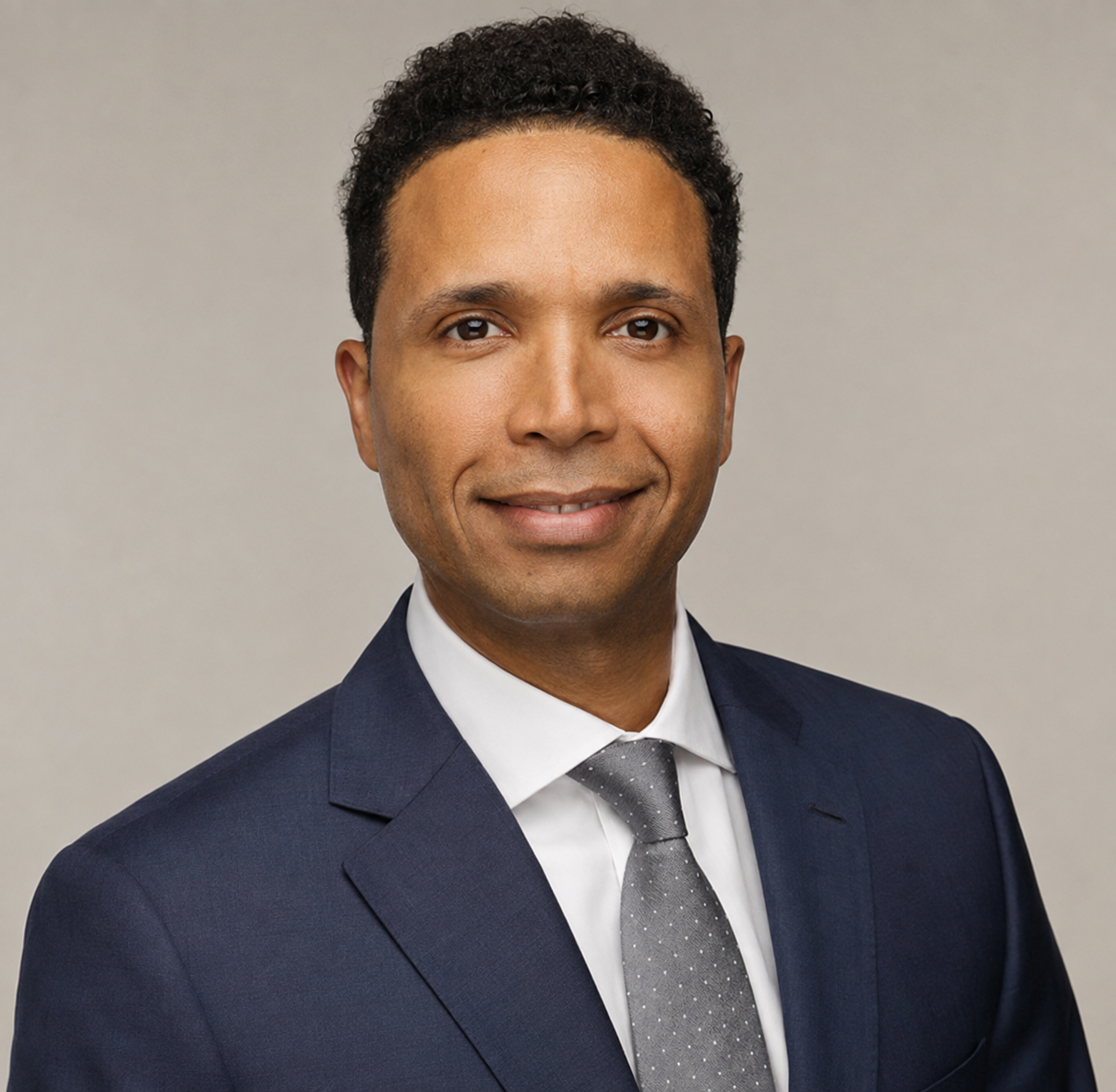 Smiling man with curly hair wearing a navy suit, white shirt, and gray polka dot tie on beige background.