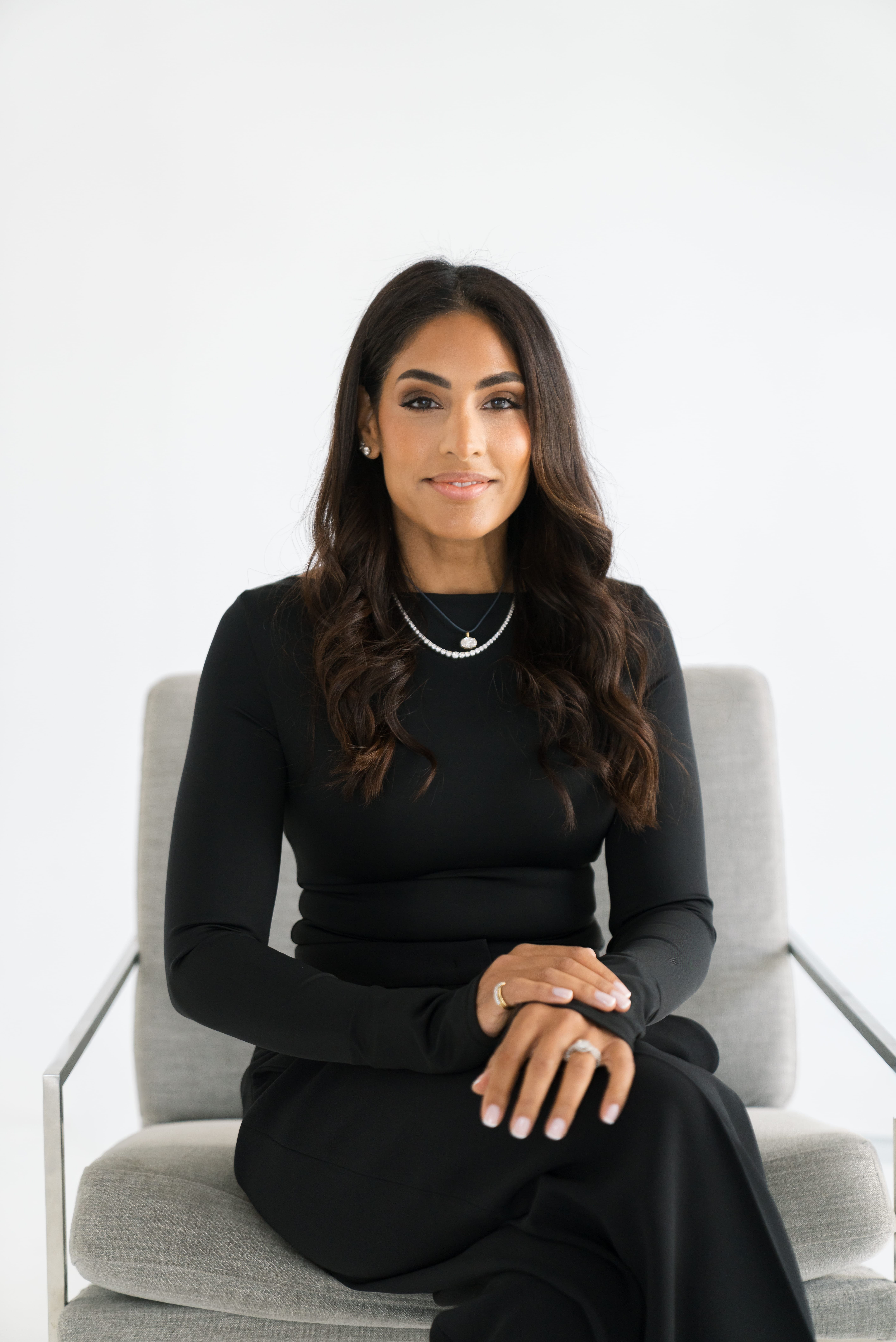 Woman with long dark hair wearing a black long-sleeve top and black pants sits on a gray armchair against a white background, with hands folded on her knee, wearing rings and necklaces, smiling softly.