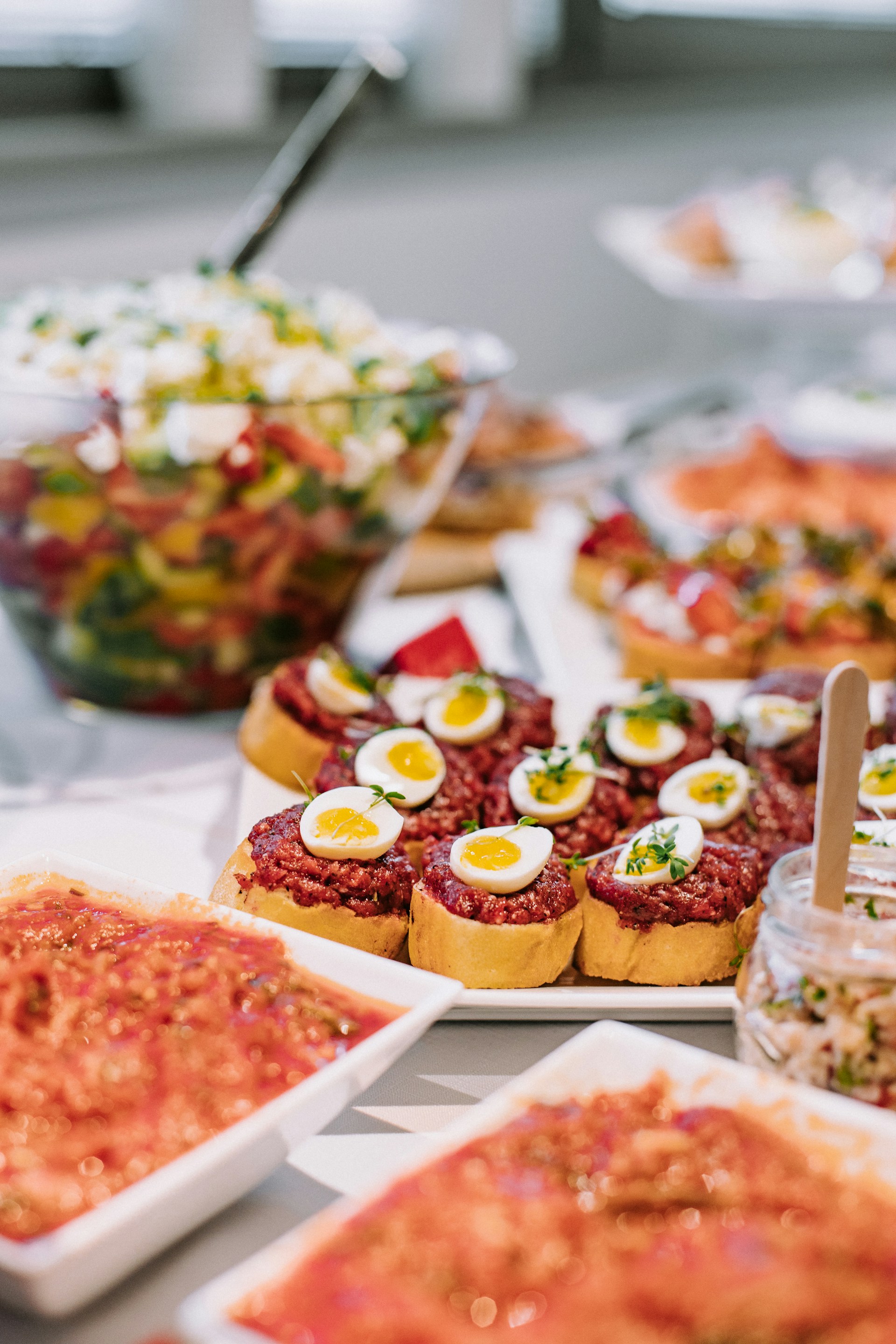 Plate of bite-sized tartlets topped with minced meat and halved boiled quail eggs garnished with herbs on a table with various dishes and salads.