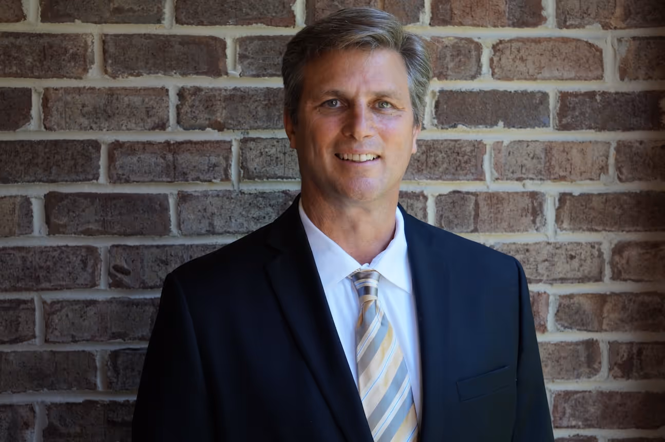 Smiling middle-aged man in a black suit, white shirt, and striped tie standing in front of a brick wall.