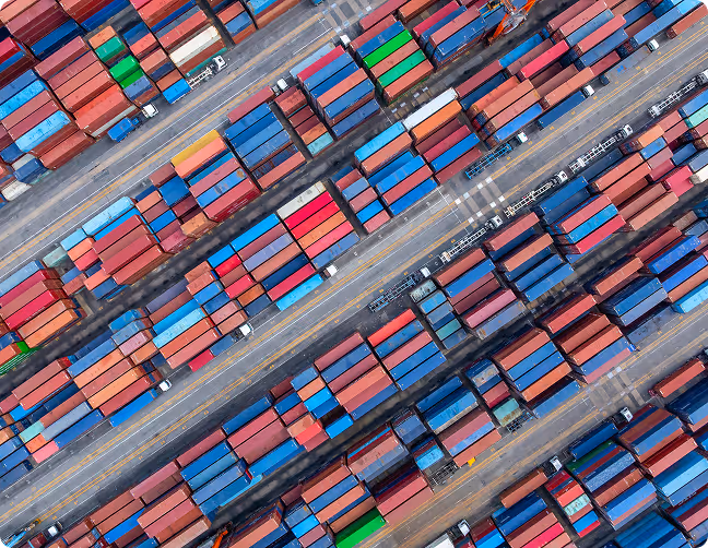 Orange excavator loading long logs into a large pile next to a purple logging truck.
