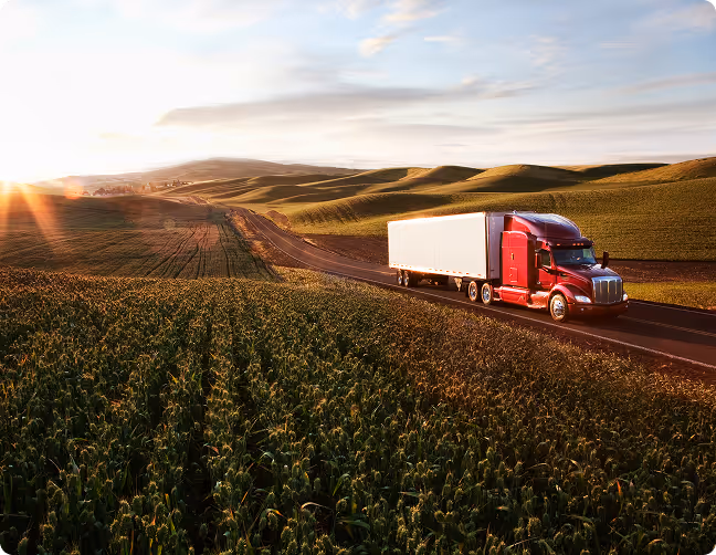 Red semi-truck with white trailer driving on a road through green farmland at sunset.