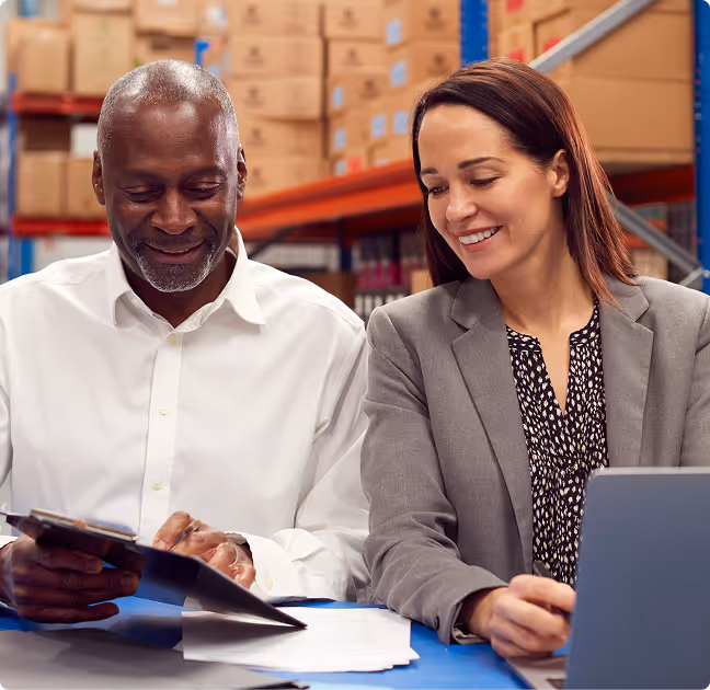 Smiling man and woman reviewing documents together at a table in a warehouse with shelves of boxes in the background.