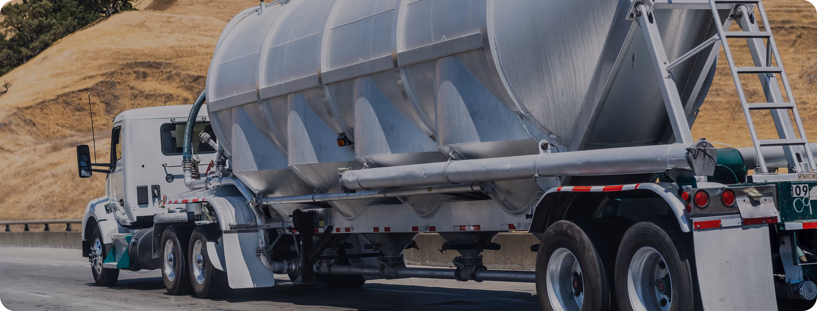 White tanker truck driving on a highway with dry grassy hills in the background.