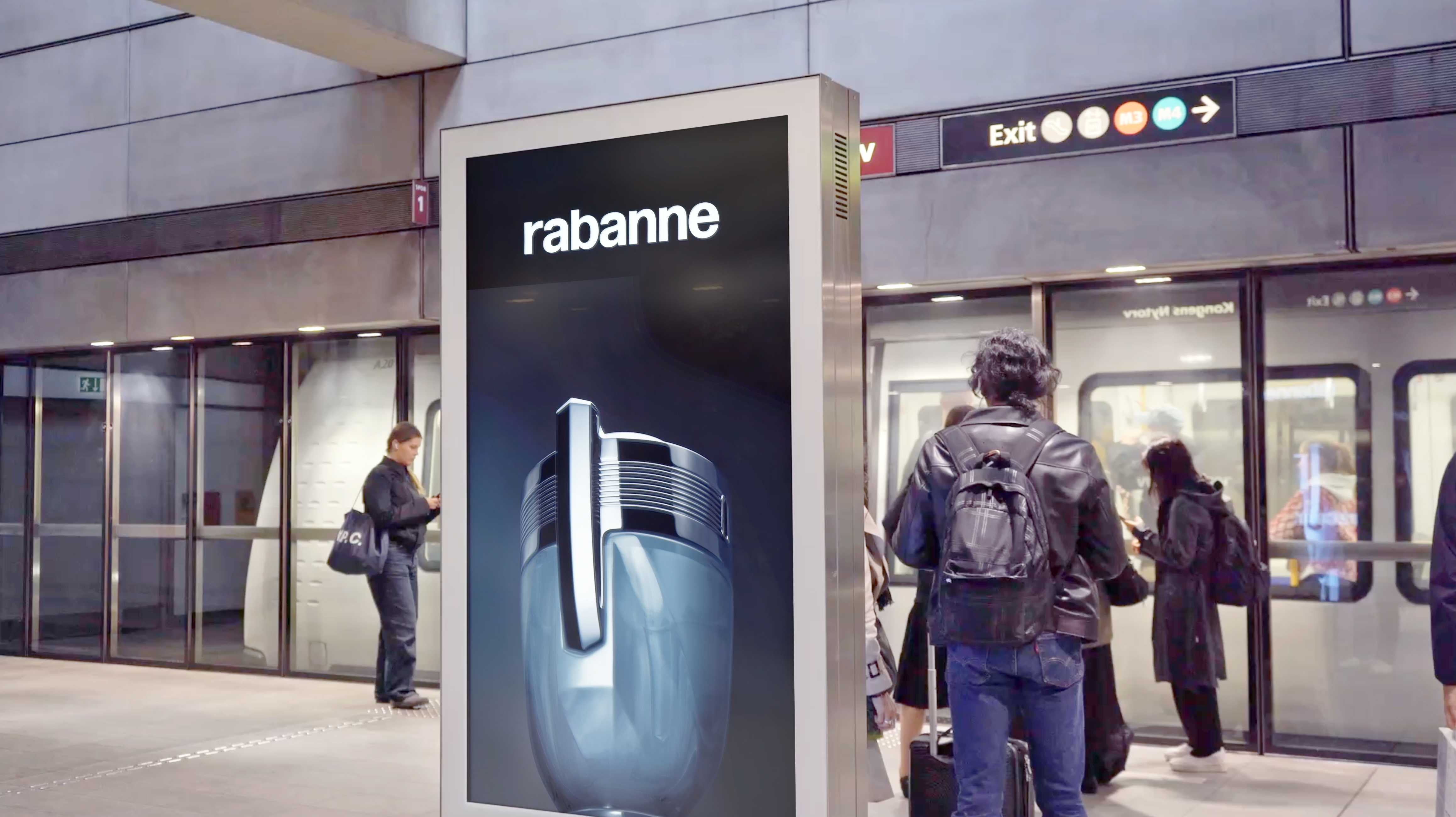 På billedet ses en metrostation, hvor passagerer enten venter på eller er i færd med at stige ombord på toget. En digital billboard viser en reklame for Rabanne, sandsynligvis for en parfume eller cologne, med en tydelig visning af produktets flakon. I baggrunden ses metroperronen, glasdøre og et skilt med retningsangivelser, hvor forskellige farvede cirkler repræsenterer de forskellige metrolinjer.