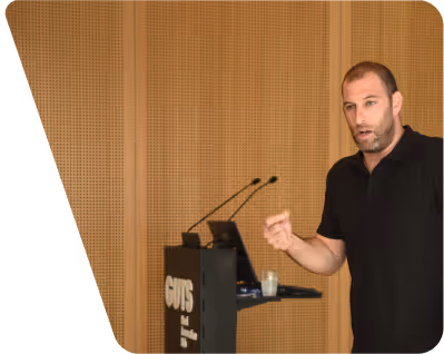 Man in black shirt speaking and gesturing near a podium with two microphones in a wood-paneled room.