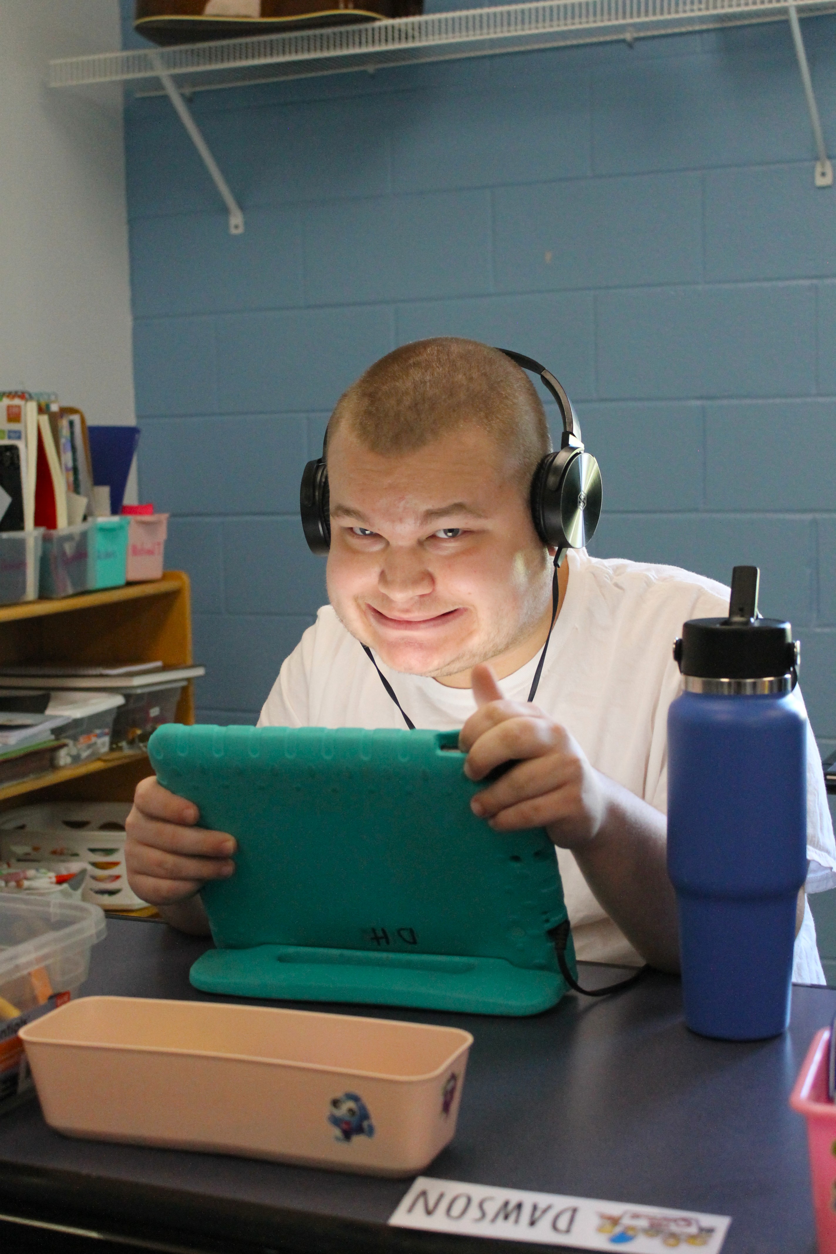 Smiling boy wearing headphones holding a green tablet at a desk with a water bottle and name tag