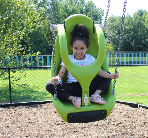 Smiling young girl sitting cross-legged in a green child safety swing at a playground.